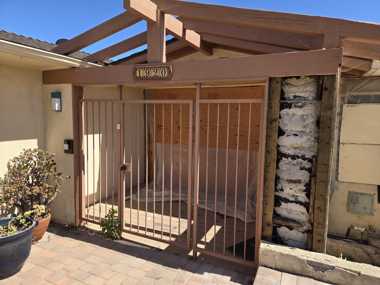 Existing Ventura front entry portico with security gate, address beam, boarded opening, and exposed right-side framing